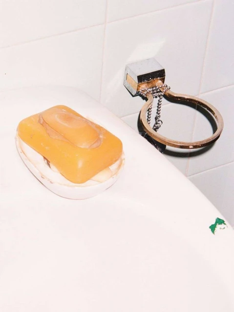 Close-up studio shot of a bar of orange soap in a white soap dish next to a metal soap holder on a tiled white wall.