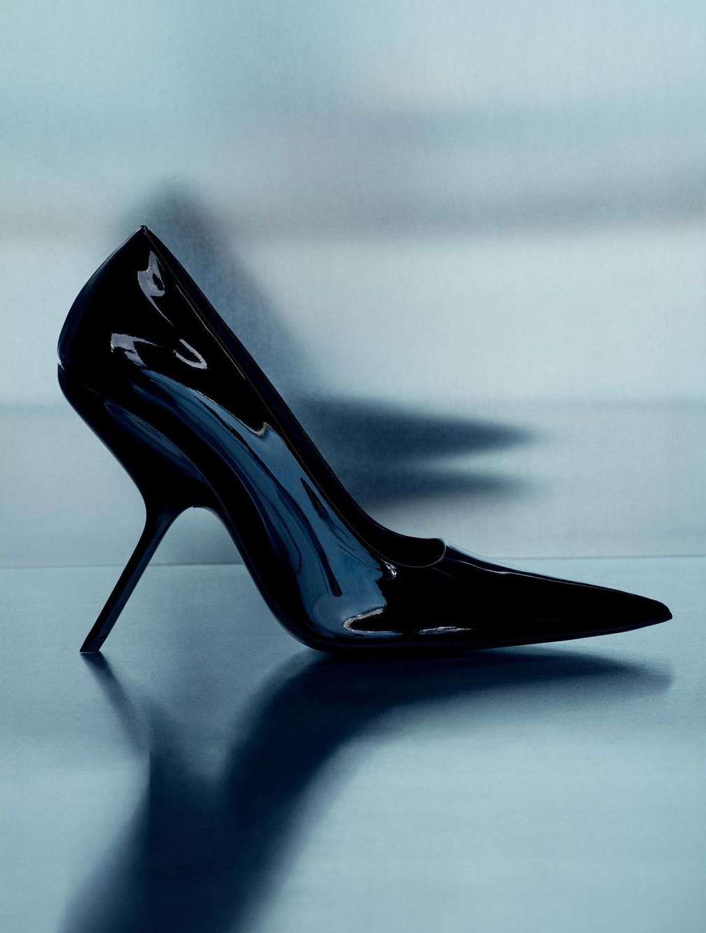 Studio shot of a glossy black stiletto heel shoe with a pointed toe, set against a blurred light blue gradient background.