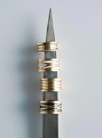 Close-up of four stacked gold rings with varying designs on a metal display stand against a plain white background, jewellery product.