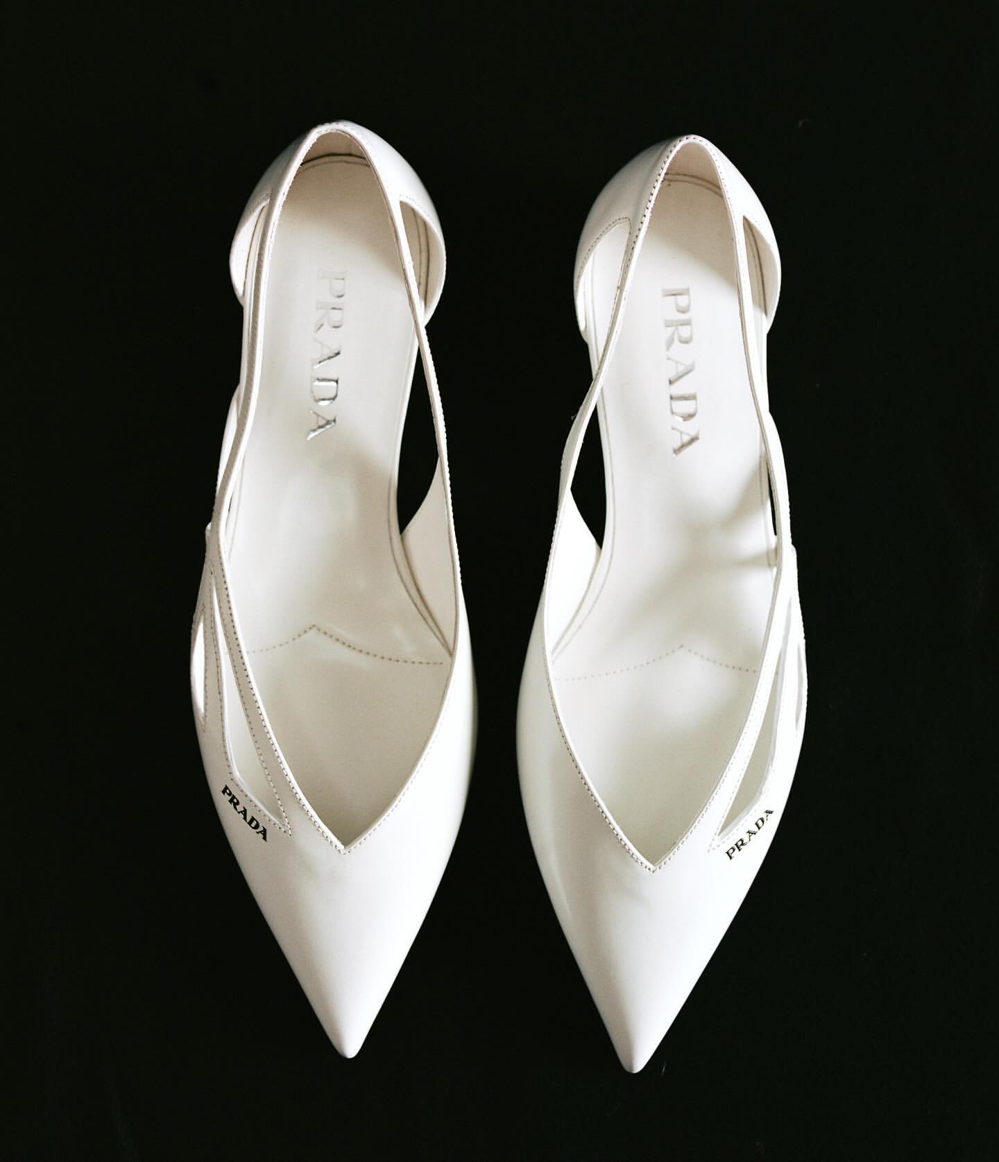 Pair of white Prada heels, pointed toe, graphic cut-outs, logo print, on black backdrop; luxury shoes, fashion editorial, product photography.