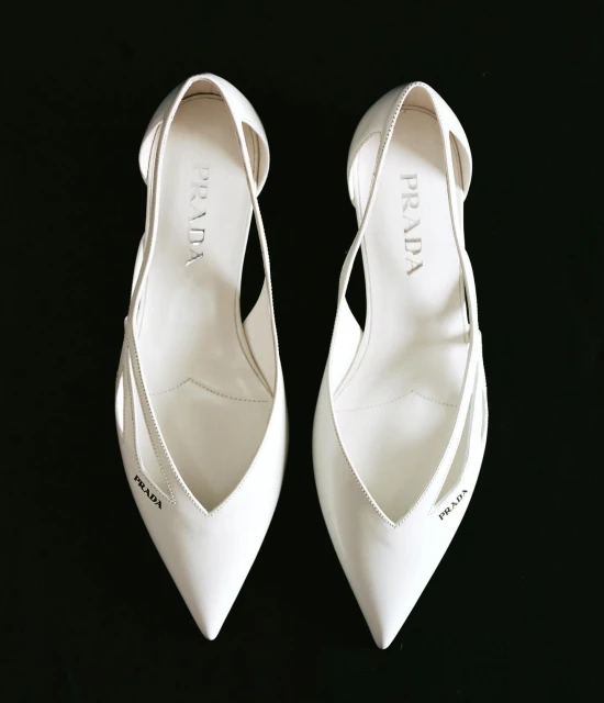 Pair of white Prada heels, pointed toe, graphic cut-outs, logo print, on black backdrop; luxury shoes, fashion editorial, product photography.