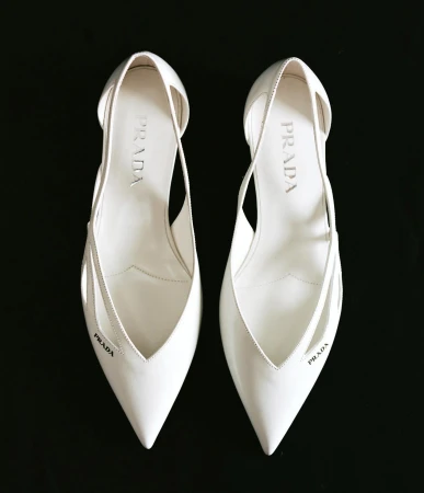 Pair of white Prada heels, pointed toe, graphic cut-outs, logo print, on black backdrop; luxury shoes, fashion editorial, product photography.