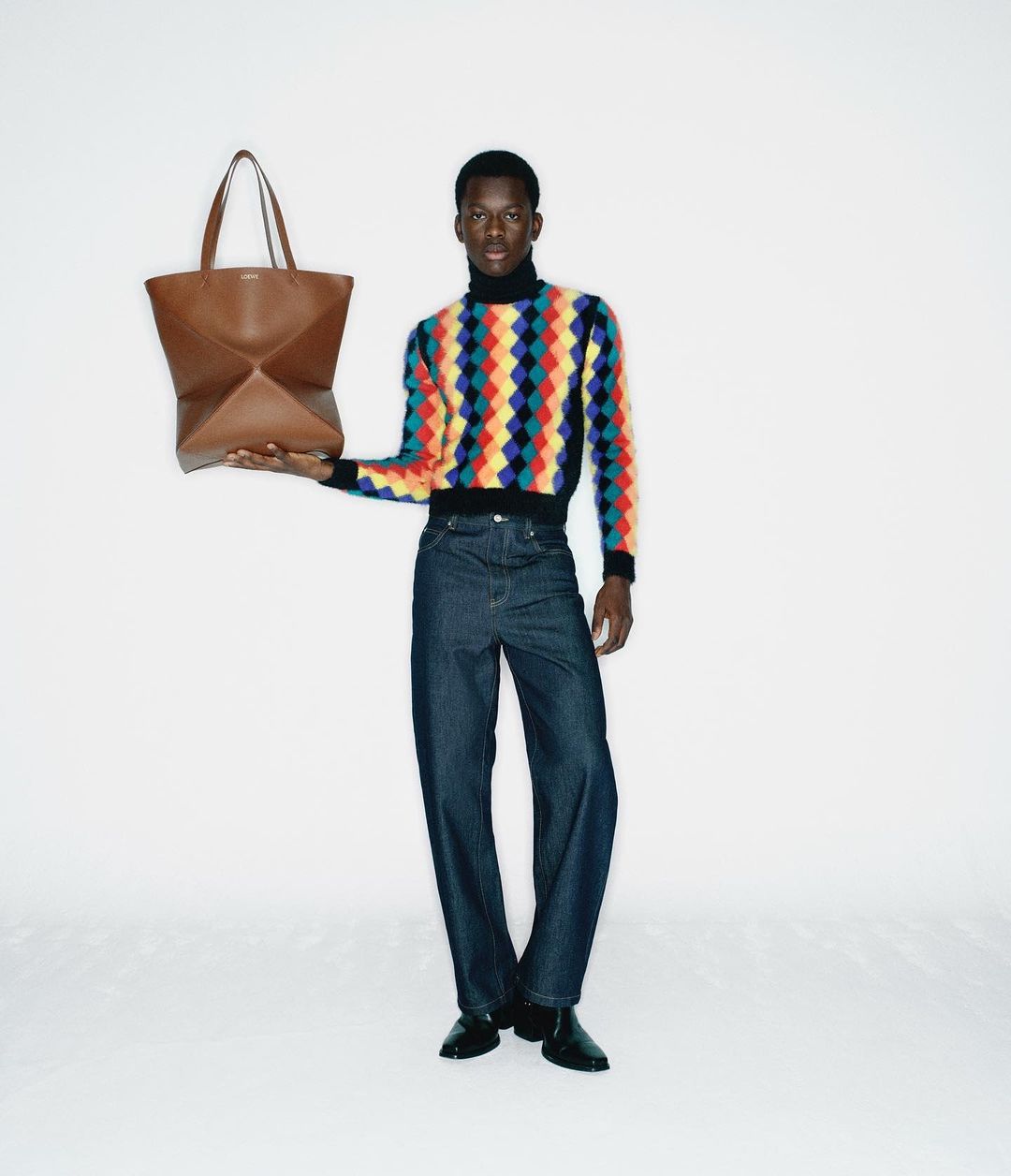 Black man holding a brown leather tote bag, wearing a colorful patterned turtleneck and dark jeans