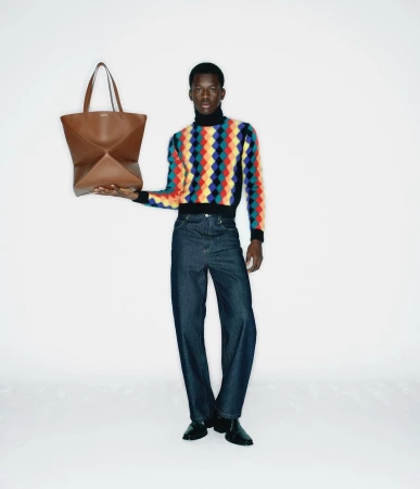 Black man holding a brown leather tote bag, wearing a colorful patterned turtleneck and dark jeans