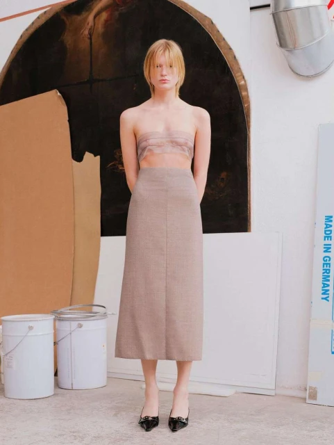 A full portrait of a slender fashion model with blonde hair posing in a taupe skirt and tan top against a painterly studio backdrop.