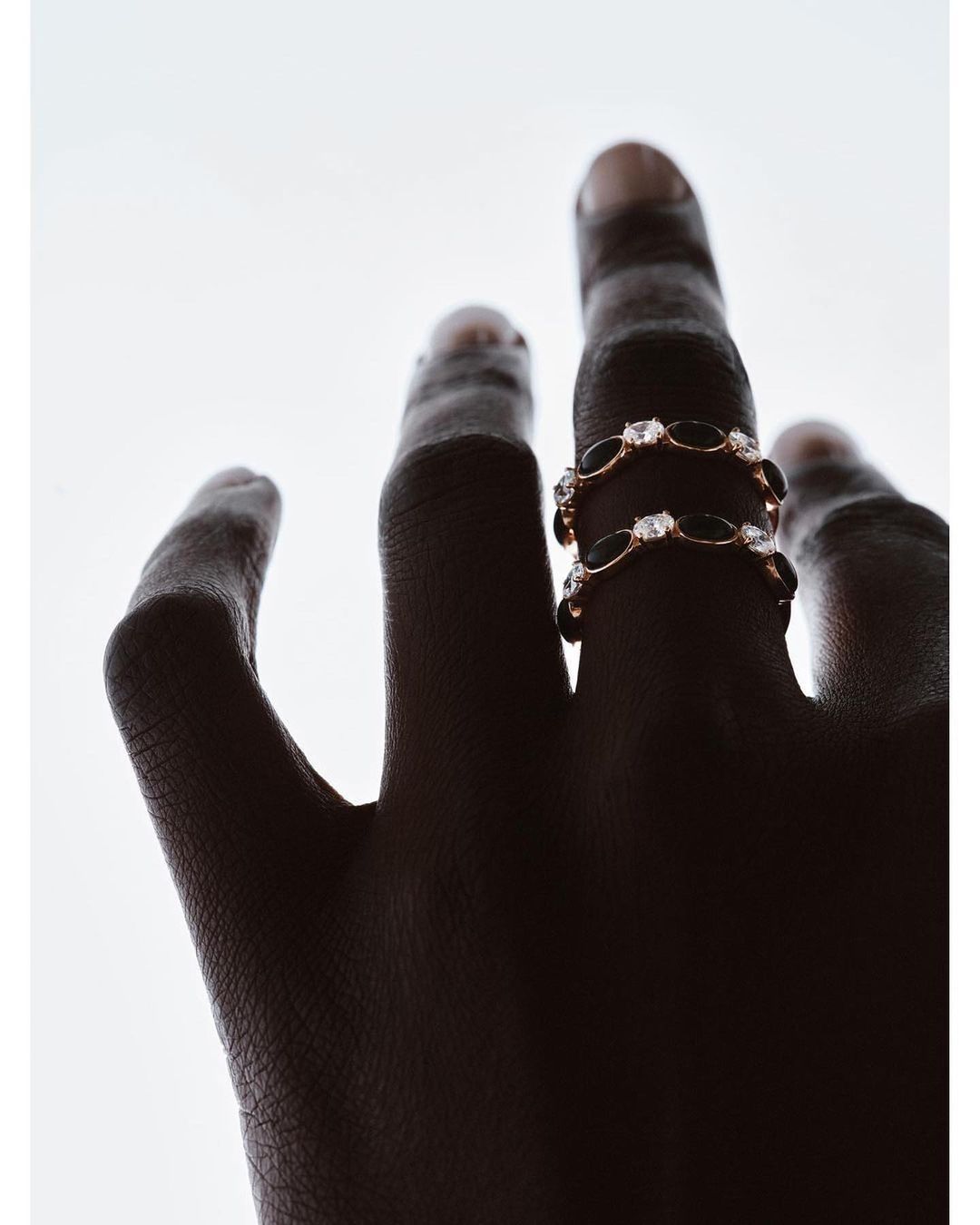 Hand with dark skin wearing two gold rings adorned with black and clear gemstones, set against a bright white backdrop.