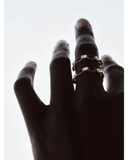 Hand with dark skin wearing two gold rings adorned with black and clear gemstones, set against a bright white backdrop.