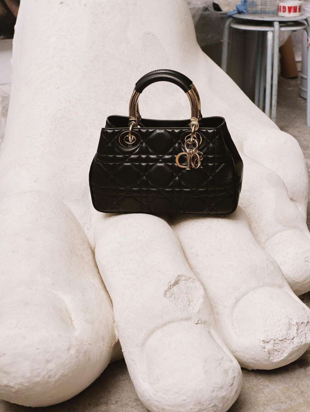 Black Dior handbag with gold hardware sits on a large white hand sculpture in a neutral indoor setting, showcasing high-fashion contrast.