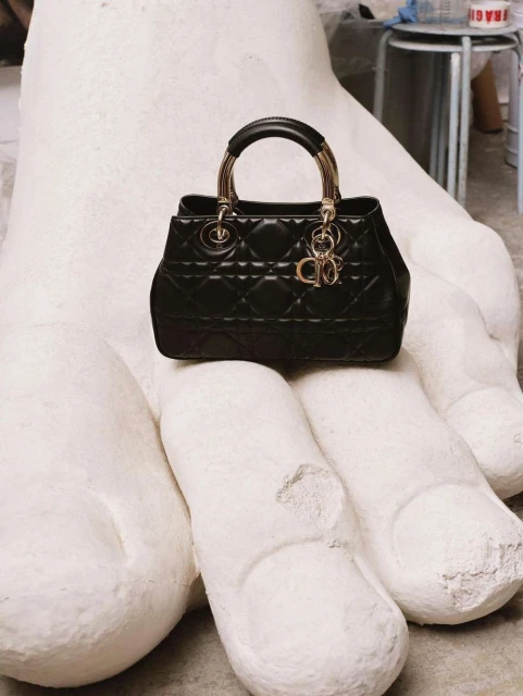 Black Dior handbag with gold hardware sits on a large white hand sculpture in a neutral indoor setting, showcasing high-fashion contrast.