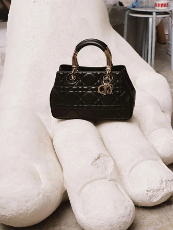 Black Dior handbag with gold hardware sits on a large white hand sculpture in a neutral indoor setting, showcasing high-fashion contrast.