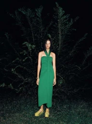 Woman wearing a long green halter dress and platform sandals standing outdoors at night in front of dark foliage
