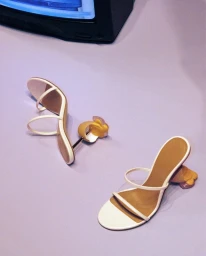 High-angle view of two white high-heeled sandals with sculptural heels resting on a pale purple surface near a television screen
