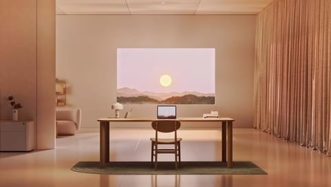 Minimalist interior with a wooden desk, chair, and a laptop under a landscape painting, bathed in warm, soft lighting.