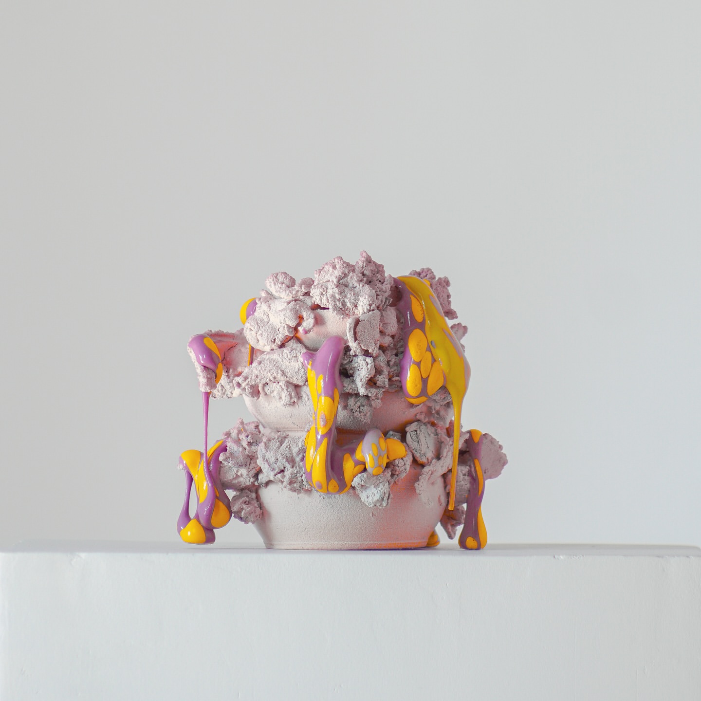 Abstract sculpture with a rough texture, featuring purple and yellow drips against a plain background, creating a studio art piece.