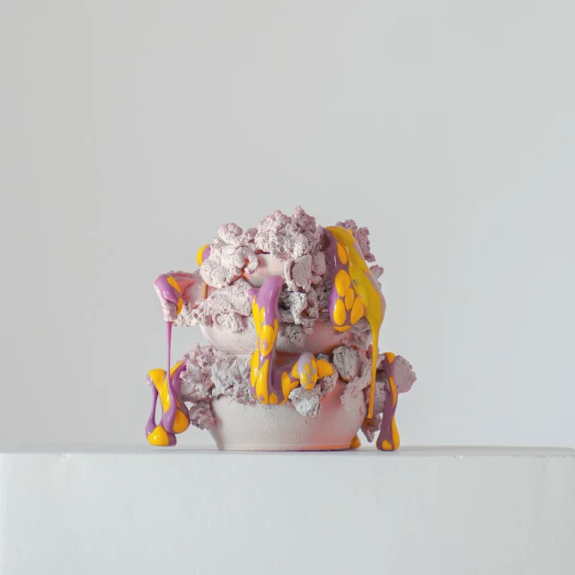 Abstract sculpture with a rough texture, featuring purple and yellow drips against a plain background, creating a studio art piece.