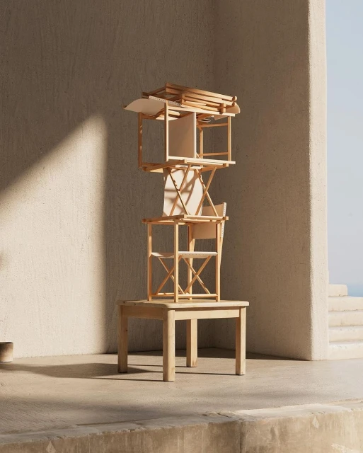 Abstract art installation with a stacked arrangement of wooden tables and geometric objects in a sunlit interior space.