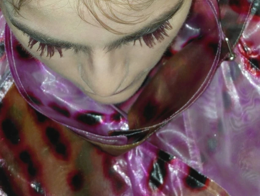 Close-up on a person's face with visible dark false eyelashes, fair skin, and a transparent raincoat with a pink and brown pattern.