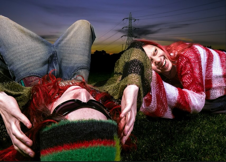 Two red-haired women laugh on green grass beneath power lines at dusk, one woman lying down with jeans, the other leaning in a striped sweater.