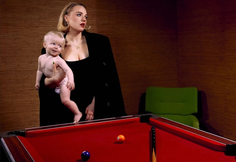 Woman in a black blazer holds an infant next to a billiards table in an indoor setting with wood-paneled walls and a green chair.