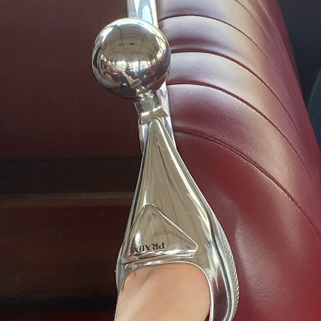 Close-up of a person wearing a silver Prada sandal, sitting on a vintage car seat with red upholstery and a silver gear stick.