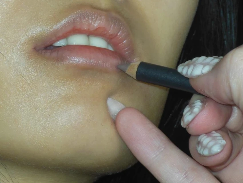 Close-up shot of a person applying lip liner with a pencil. The focus is on the lips and the hand holding the cosmetic pencil.