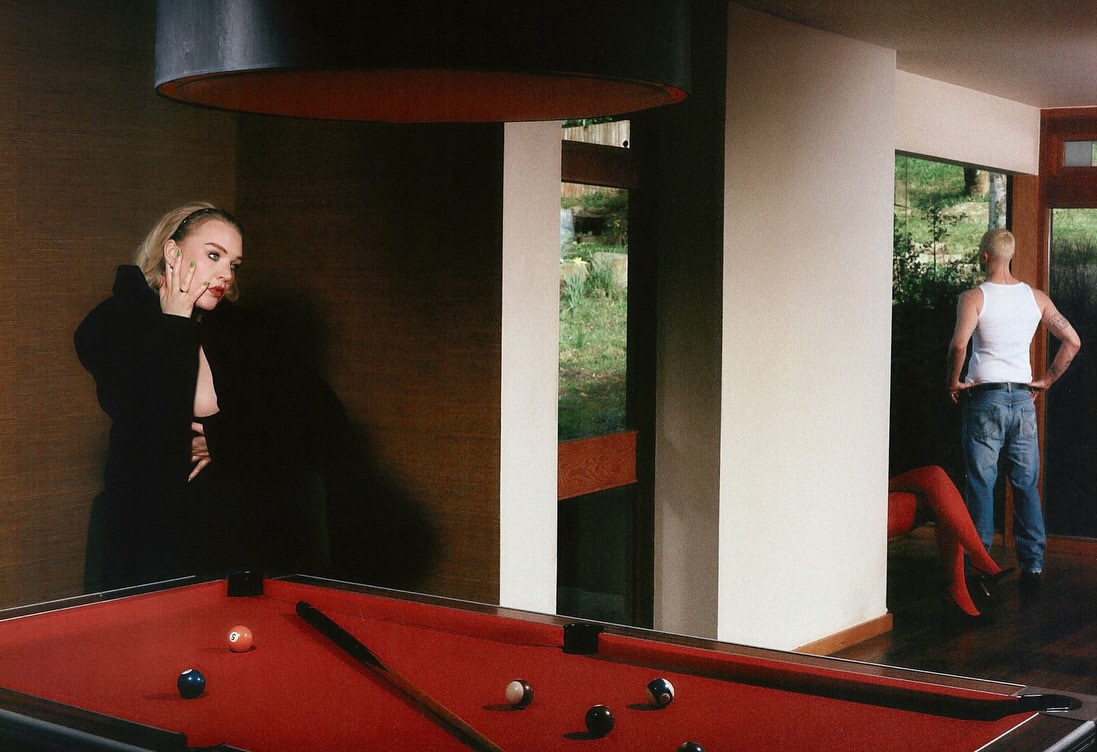 Woman in black near red pool table indoors, man with white tank top stands in background, bathed in natural light.