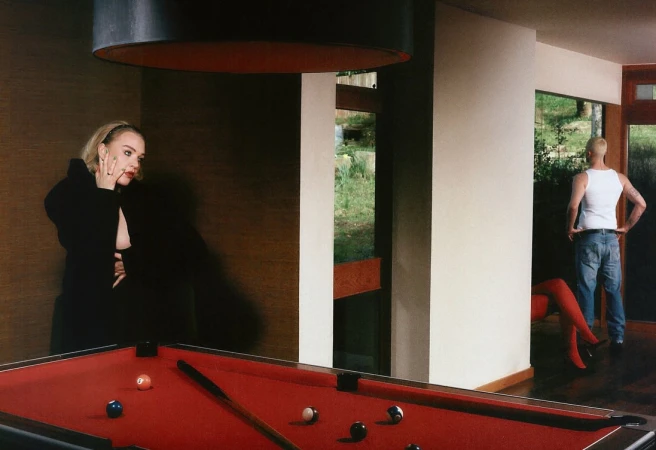 Woman in black near red pool table indoors, man with white tank top stands in background, bathed in natural light.
