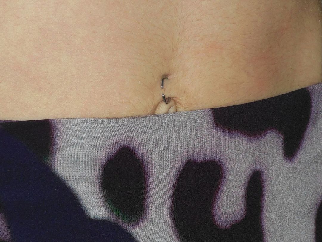 Close-up of an abdomen showing a navel piercing and a blurred abstract pattern on leggings. Skin tone is fair with visible body hair.