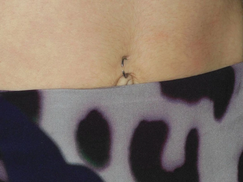 Close-up of an abdomen showing a navel piercing and a blurred abstract pattern on leggings. Skin tone is fair with visible body hair.