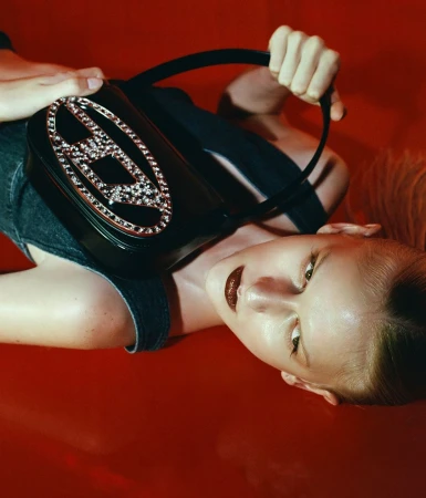 Fashion shot of a blonde woman lying on a red surface, holding a black purse with a silver emblem, glossy lips, and wearing a denim top.