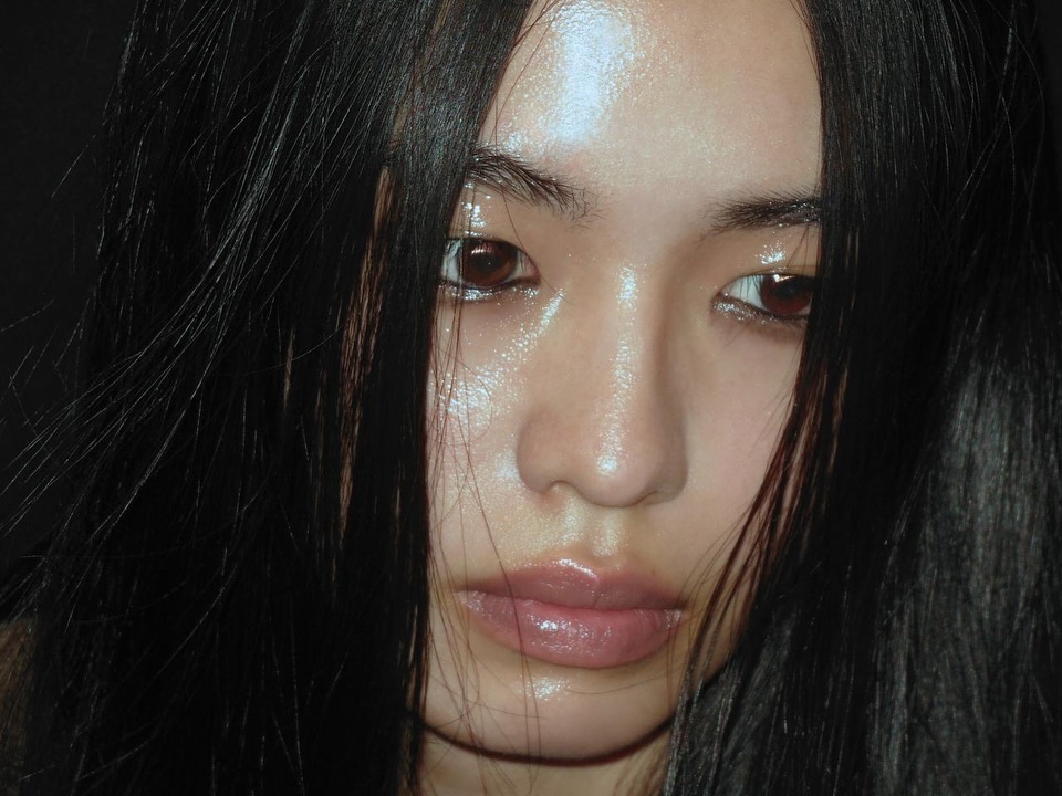 Close-up portrait of a woman with dark hair and a glossy complexion, gazing directly at the camera against a dark background.