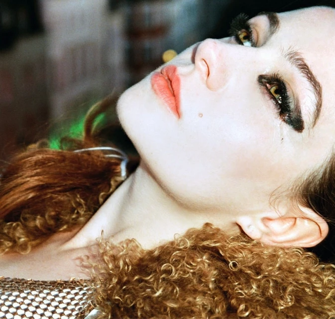 Close-up portrait of a woman with brown curly hair, wearing heavy eye makeup, with her head tilted back and lit with studio lighting.
