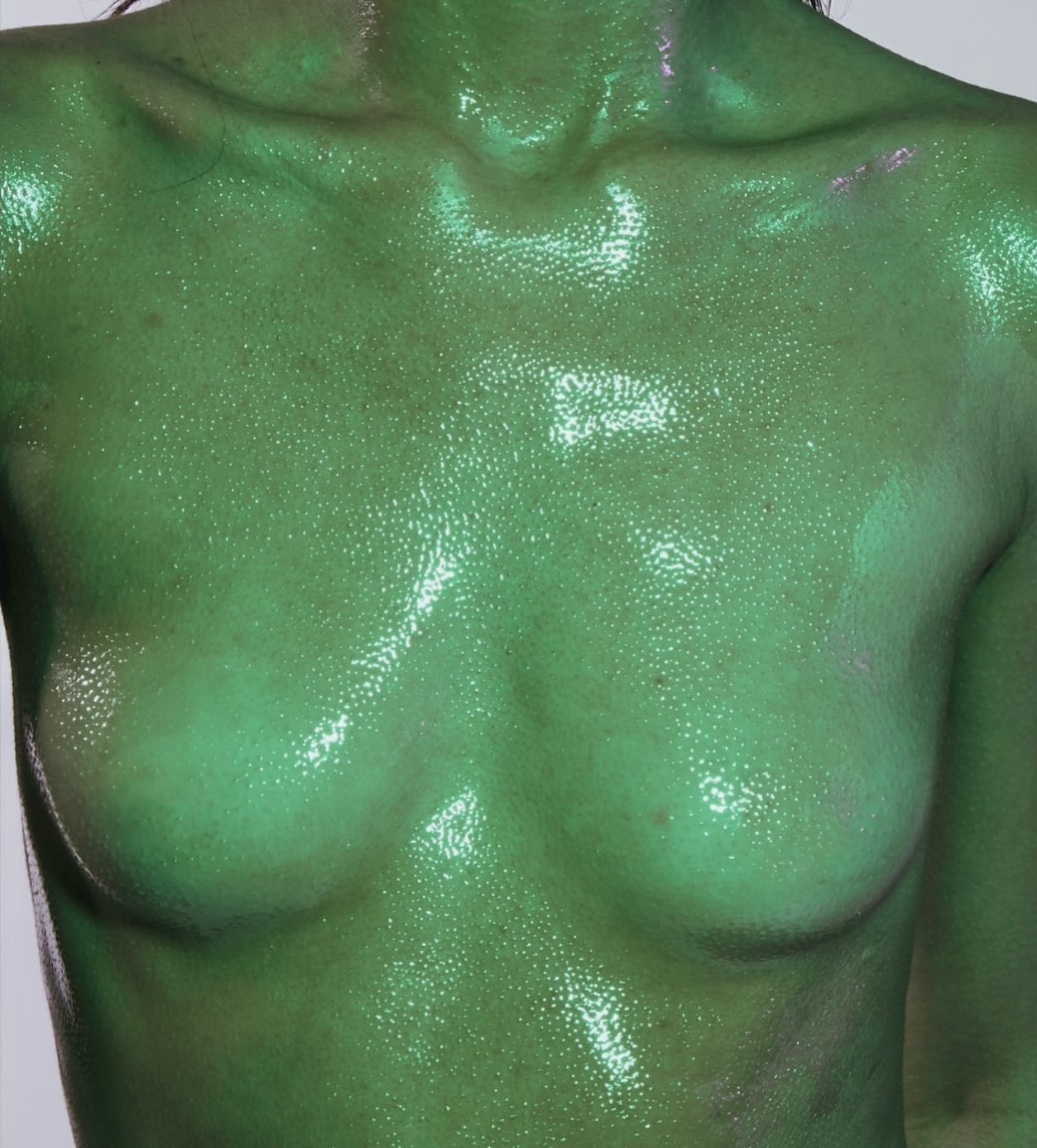 Torso of a female adult covered in a green, shimmery substance against a plain light background in a close-up shot.