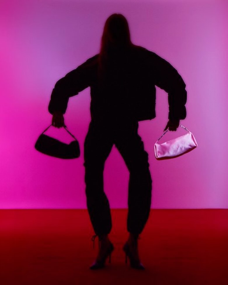 Silhouette of an adult female fashion model in a photo studio, wearing a jacket, pants, and high heels, and holding two handbags.