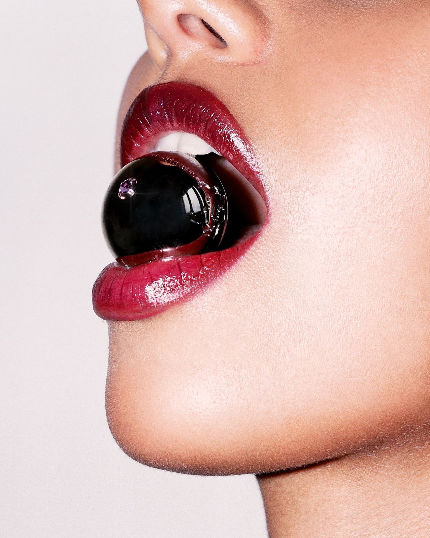 Close-up shot of a woman's face with glossy red lips holding a black gumball in her mouth against a light grey background.