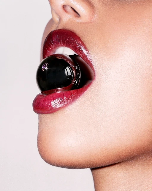 Close-up shot of a woman's face with glossy red lips holding a black gumball in her mouth against a light grey background.