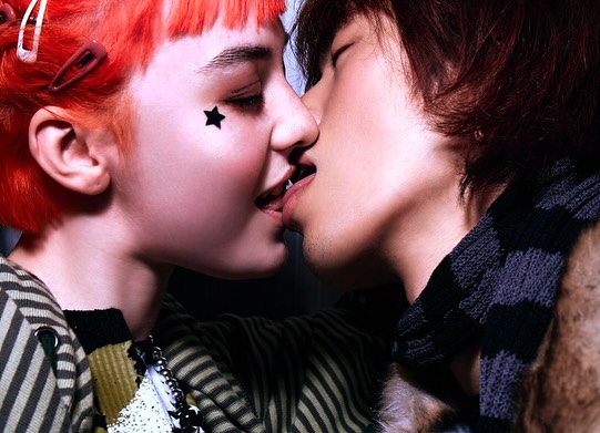 Close-up of a young adult couple kissing, with visible skin tones and details like hair clips, makeup, and striped clothing textures.