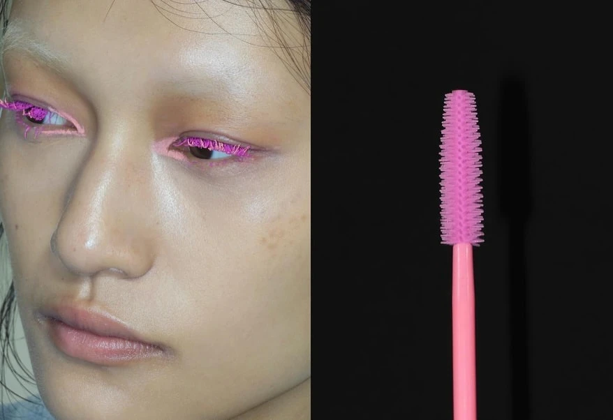 Close-up of an adult female with pink mascara on her eyelashes alongside a pink mascara wand against a black background.