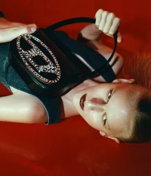 Model lying on a glossy red surface holding a black shoulder bag with a rhinestone logo by Yana Van Nuffel