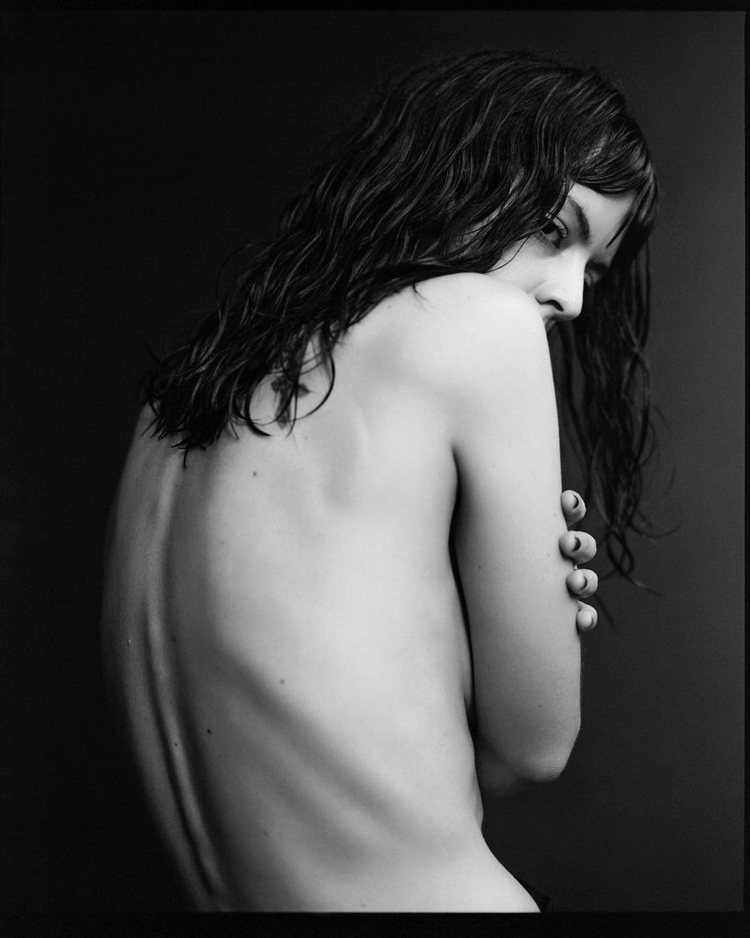 Black and white portrait of a slender adult woman with long dark hair, bare back, arms crossed, against a dark grey backdrop.
