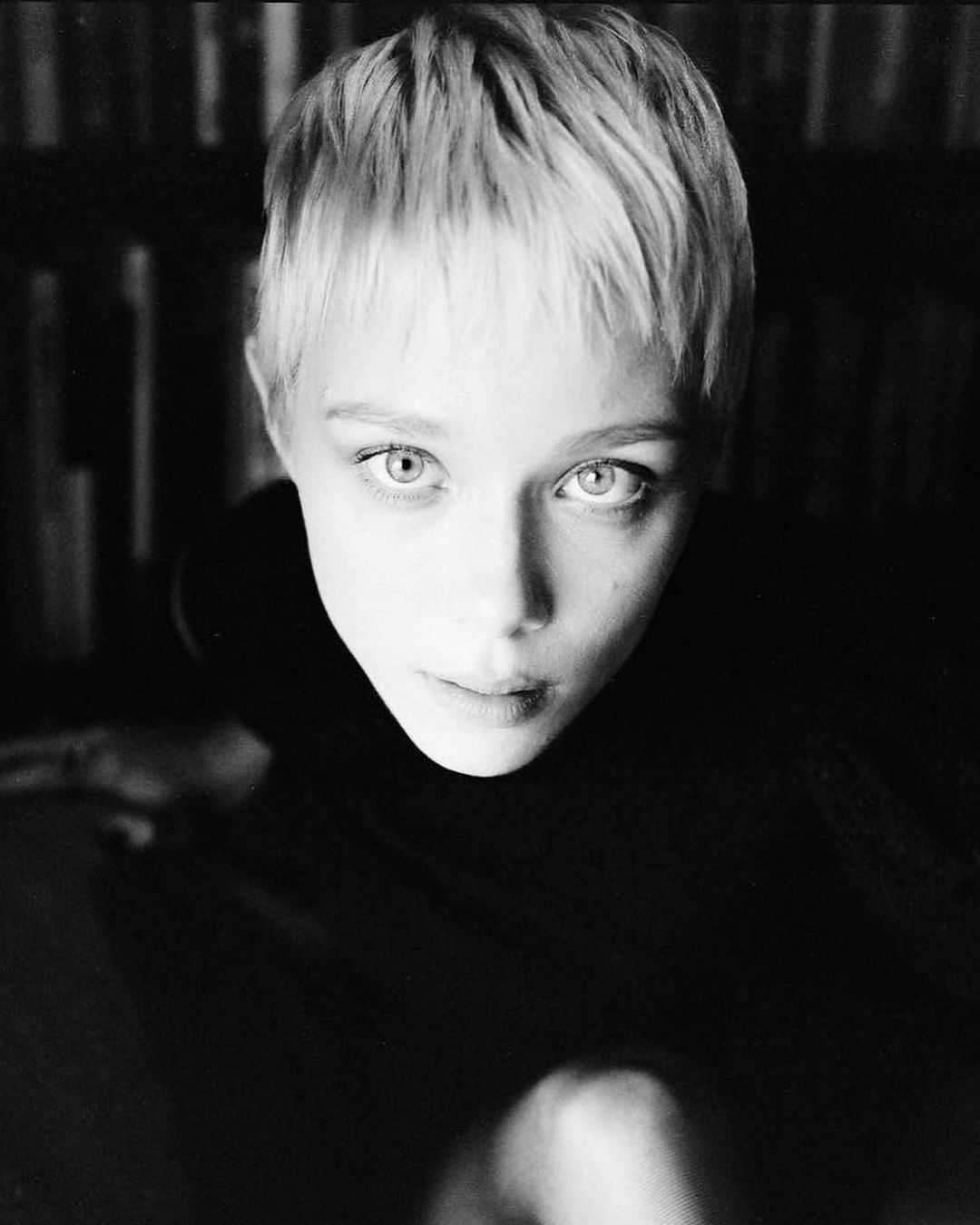 Striking black and white portrait of a young adult with short blonde hair, shot from a high angle in an indoor environment with dark background.