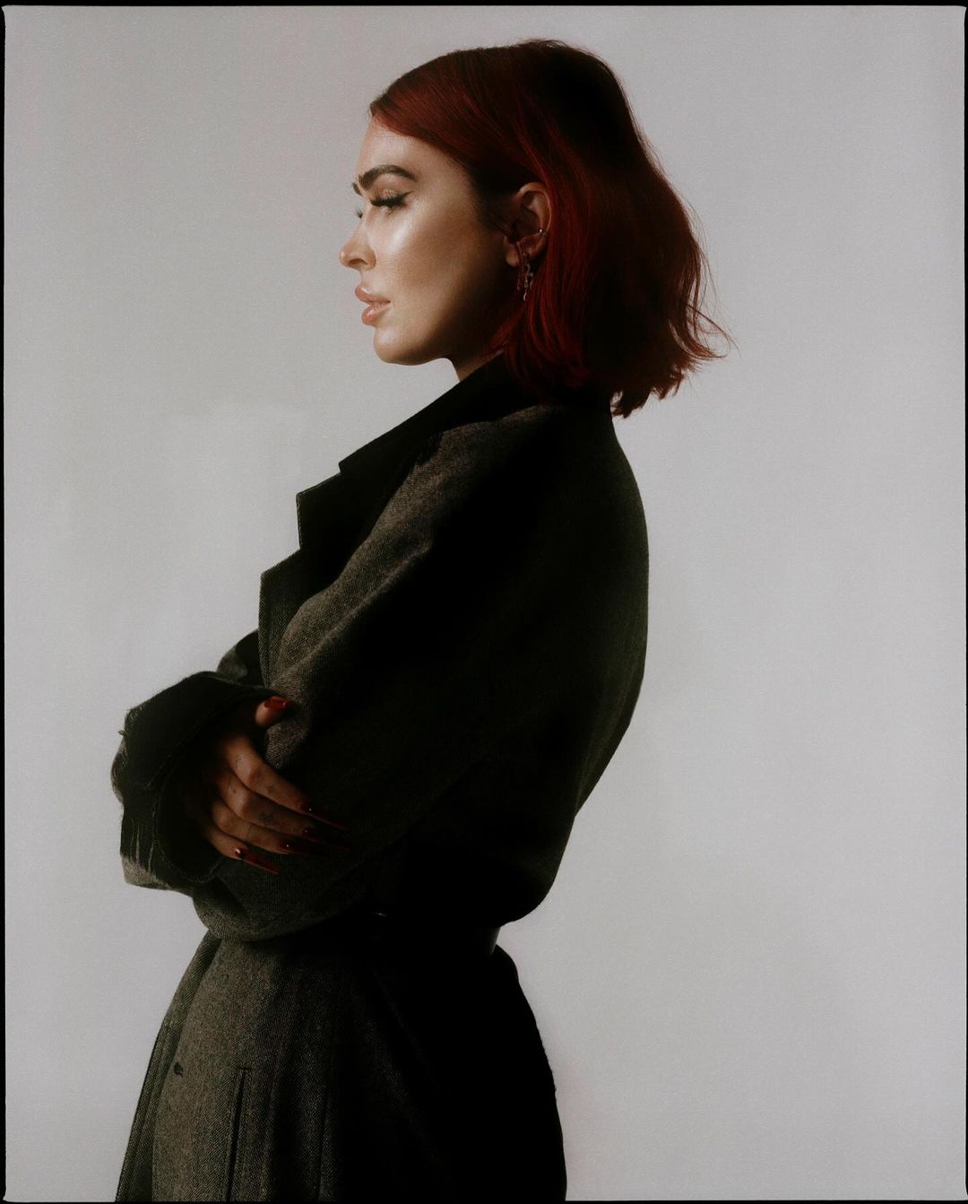 Profile portrait of a red-haired woman with arms crossed wearing a dark coat against a plain background