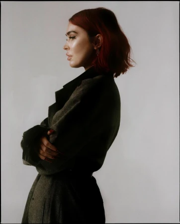 Profile portrait of a red-haired woman with arms crossed wearing a dark coat against a plain background