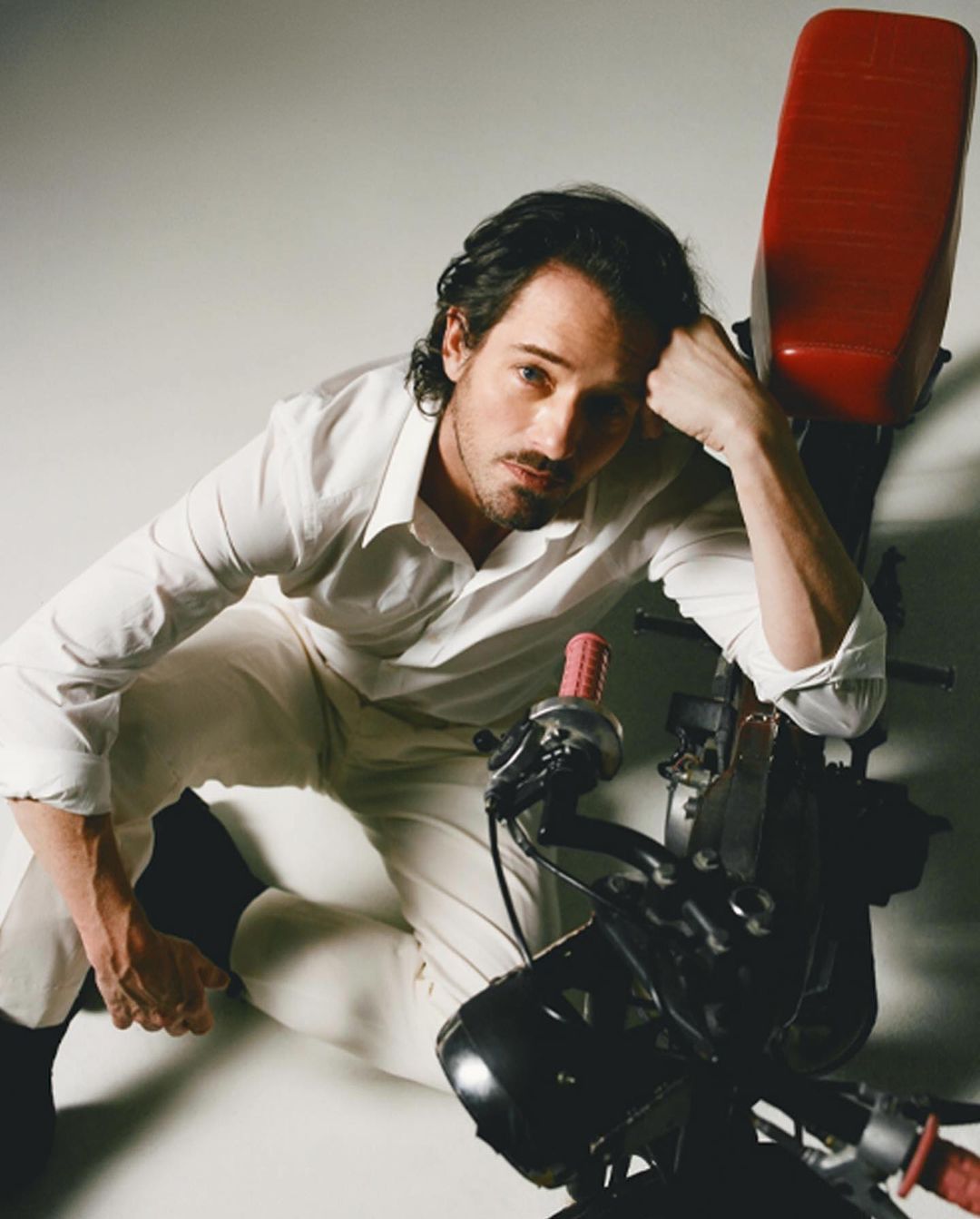 A man in a white shirt and pants poses with a black and red motorcycle in a studio setting, showcasing fashion and automotive elements.