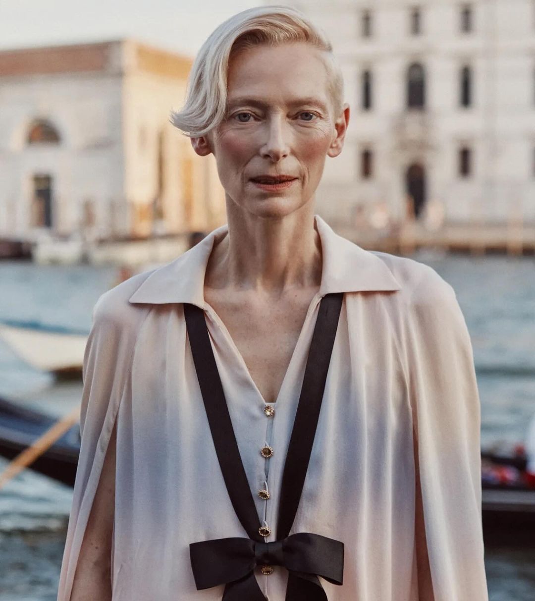 Elegant woman with short blonde hair wearing a light pink dress with a black bow, posing in front of a canal and building in Venice.