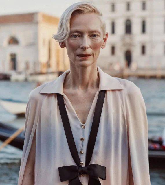 Elegant woman with short blonde hair wearing a light pink dress with a black bow, posing in front of a canal and building in Venice.