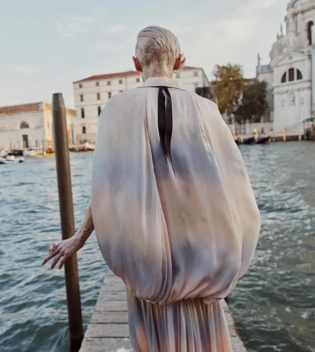 A woman stands on a dock near the water in Venice, Italy, wearing a flowing pastel dress with her back facing the camera.