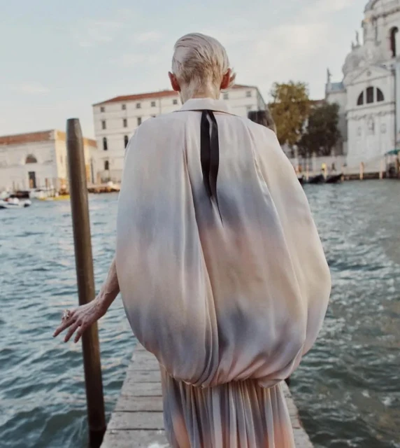 A woman stands on a dock near the water in Venice, Italy, wearing a flowing pastel dress with her back facing the camera.