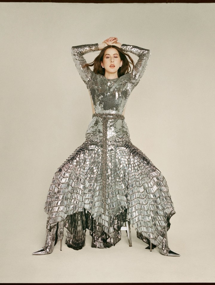 A woman with long brown hair poses in a long-sleeved silver sequin gown and matching silver boots in a photo studio setting. 