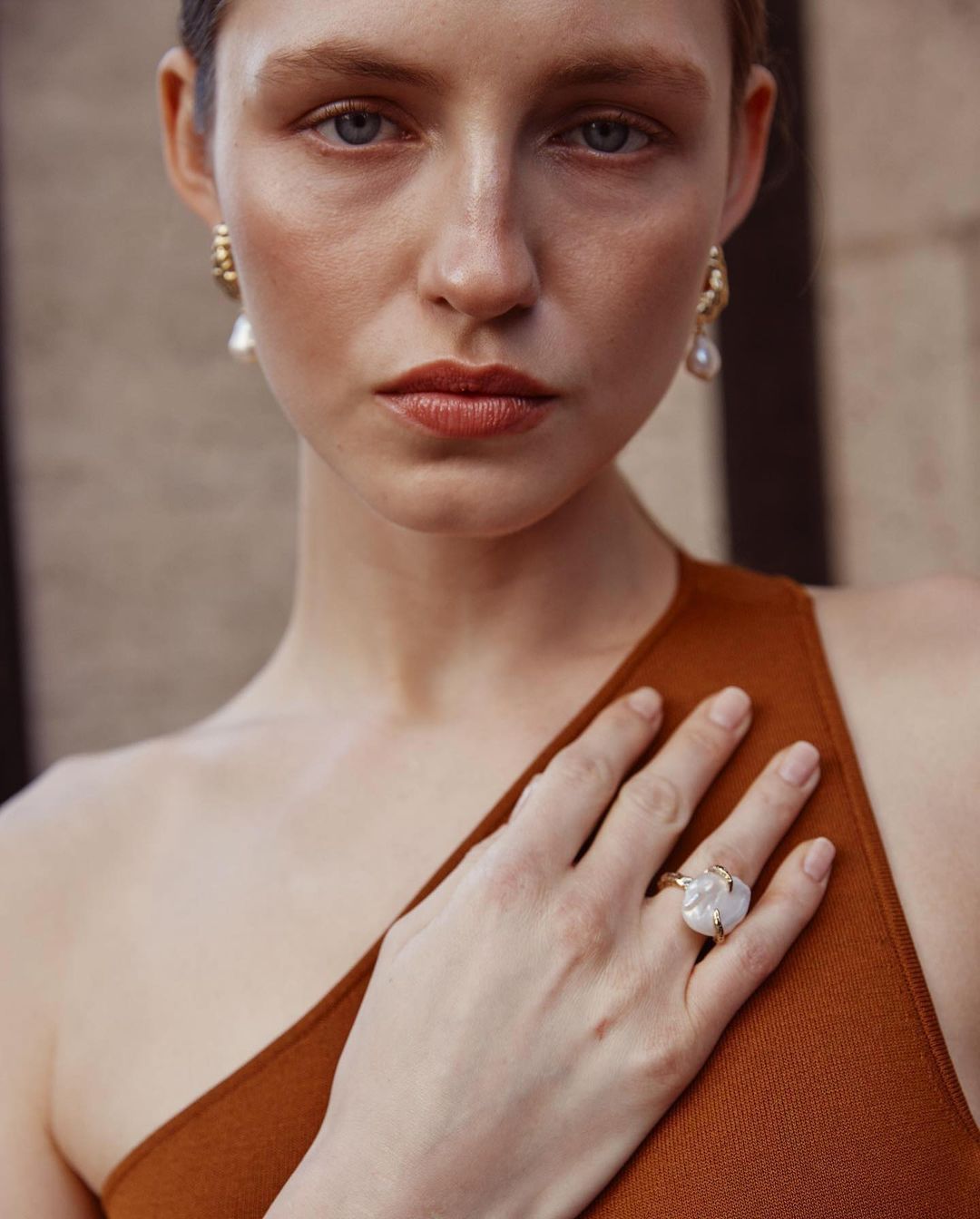 Close-up of a woman with fair skin wearing a brown dress, gold earrings, and a large ring on her right hand.
